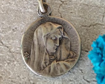 Puede incluir: Un colgante plateado con un relieve detallado de una mujer con un tocado drapeado. El colgante redondo tiene un lazo en la parte superior para una cadena. La imagen está sobre un fondo gris con un acento azul.
