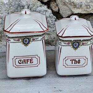 May include: Two white ceramic canisters with lids, labeled "CAFÉ" and "Thé" in red script. Each has a decorative floral band and red trim. The canisters are rectangular with a slightly angled top.