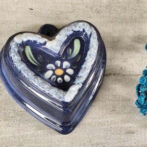 Può includere: Uno stampo in ceramica blu a forma di cuore con un design floreale bianco e verde. Lo stampo ha un bordo rialzato e un foro nella parte superiore per appenderlo.
