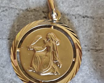 May include: Gold-colored pendant featuring a detailed relief of a seated figure holding an object, possibly a quill. The pendant has a circular design with a textured outer ring and a loop for a chain. The pendant is likely made of gold.