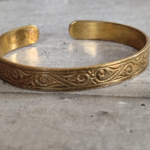 Peut inclure: Un bracelet manchette de couleur or avec un design ouvert. Le bracelet présente un motif floral complexe et répétitif autour du bracelet. Le métal a un aspect légèrement vieilli, lui donnant une esthétique vintage. Bijoux.