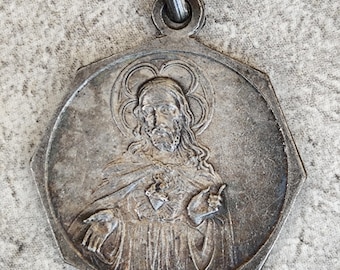 May include: A silver-toned metal pendant in a hexagonal shape featuring a raised relief of a figure with long hair and a halo. The figure has a heart emblem on the chest and is depicted with one hand raised and the other extended outward.