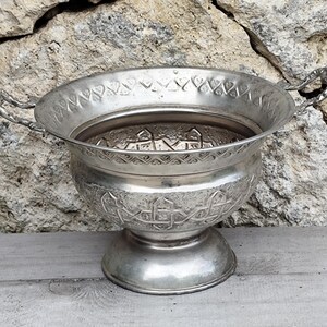 Op de afbeelding: Een zilverkleurige metalen kom met twee handvatten en een voet. De kom heeft reliëfpatronen rond de rand en de body. De kom is waarschijnlijk voor decoratief gebruik. Diameter ongeveer 25 cm.