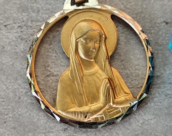 Puede incluir: Colgante dorado con una figura religiosa en oración, enmarcada en un círculo con borde facetado. El colgante tiene una argolla para sujetar a una cadena, ideal para un collar.