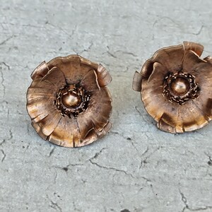 May include: A pair of copper-colored flower-shaped stud earrings. Each earring features detailed petals and a textured center, creating a realistic floral design. The earrings are displayed on a textured gray surface.