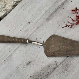 Puede incluir: Un servidor de pastel plateado vintage con un diseño floral ornamentado en la hoja. El mango tiene un patrón decorativo. El servidor se coloca sobre una superficie de madera clara y desgastada, con flores rojas en el fondo.