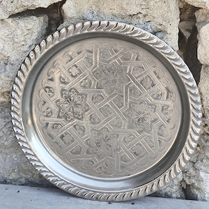 Op de afbeelding: Een rond zilverkleurig metalen dienblad met een ingewikkeld reliëfpatroon van in elkaar grijpende lijnen en bloemmotieven in het midden. Het dienblad heeft een verhoogde, dikke rand met een gedraaid touwontwerp.