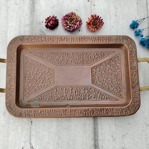 May include: A copper serving tray with two brass handles. The tray has a hammered texture and a geometric design.