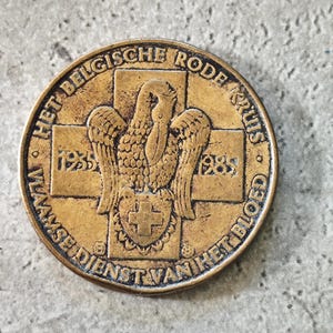 May include: A round, bronze-colored medal with text in a circular pattern around the edge. The center features a cross with a bird and shield design. The text includes "HET BELGISCHE RODE KRUIS" and "VLAAMSE DIENST VAN HET BLOED".