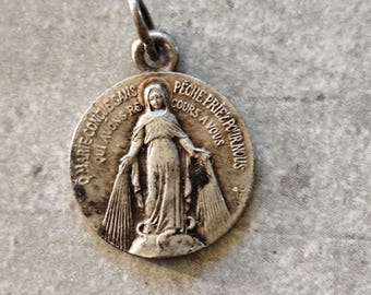 May include: A silver-toned round pendant with a loop at the top. The pendant features an embossed image of a woman with outstretched arms and text in French. The pendant is likely made of metal and has an aged appearance.