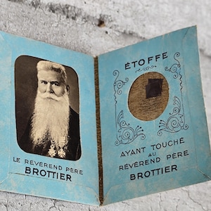 May include: A light blue paper folder featuring a black and white portrait of Le Révérend Père Brottier on the left side. The right side contains a circular cutout displaying a small piece of fabric with the text ÉTOFFE AYANT TOUCHÉ AU RÉVÉREND PÈRE BROTTIER.