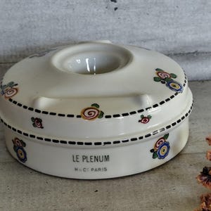 May include: Cream-colored ceramic ashtray with floral designs and the text "LE PLENUM H&C PARIS". The ashtray has a circular shape with a central opening and a dotted black border. Decorative flowers in red, blue, and yellow.