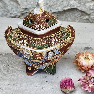 Peut inclure: Un brûleur d'encens décoratif en céramique orné d'un couvercle. Le brûleur est principalement marron avec des accents dorés, verts et blancs. Il présente un motif floral et figuratif détaillé. Le brûleur a trois petits pieds et un petit fleuron blanc sur le couvercle.