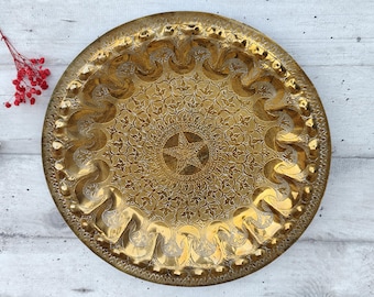 May include: A large, round, gold-colored metal tray with an intricate, embossed design. The tray has a raised rim and a center design that resembles a star.