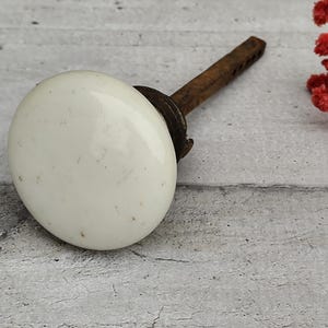 Puede incluir: Un pomo de puerta de porcelana blanca con un vástago de metal oxidado. El pomo es redondo y tiene una superficie ligeramente texturizada.