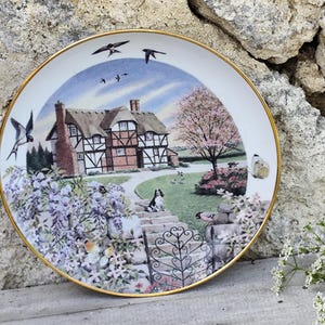 May include: Decorative plate with a gold rim featuring a detailed illustration of a cottage with a thatched roof, surrounded by a garden with flowers, butterflies, and birds. A dog sits on the steps leading to the cottage.