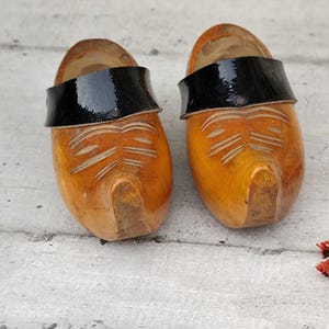 Puede incluir: Un par de zuecos de madera con acabado de madera natural y correas de cuero negro. Los zuecos tienen detalles tallados en la puntera y se exhiben sobre una superficie gris claro. Una ramita de flores secas está en el fondo.