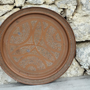 May include: A round, copper-colored tray with an intricate, embossed floral design. The tray has a raised edge and a detailed pattern in the center. The background is a rough stone wall.