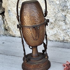 May include: Antique copper kettle with a hammered finish and a handle. The kettle sits on a decorative stand with three legs.