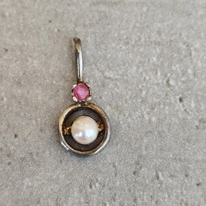 Può includere: Ciondolo in argento con una perla bianca racchiusa in un anello d'argento. Sopra l'anello c'è una piccola gemma rosa rotonda. Il ciondolo ha un piccolo anello nella parte superiore per una catena. Il gioiello è posto su uno sfondo neutro.