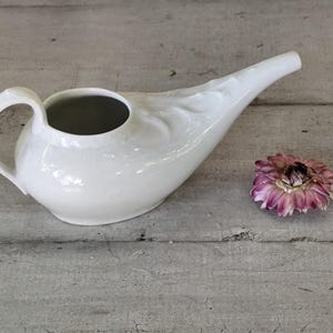May include: A white ceramic neti pot with a curved spout and handle, designed for nasal irrigation. A dried purple and pink flower rests beside the pot, adding a touch of color. The pot is on a weathered, light-colored wooden surface.