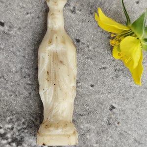 May include: A small, off-white figurine with a bottle-like shape. The figurine has a detailed, textured surface and a pedestal base. A yellow flower with green leaves is in the background. The figurine appears to be made of a light-colored material.
