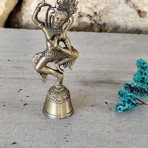 May include: A small, antique-style brass bell featuring a dancing figure. The figure is adorned with intricate details, including a headdress and ornate clothing. The bell is approximately 3 inches tall and stands on a concrete surface.