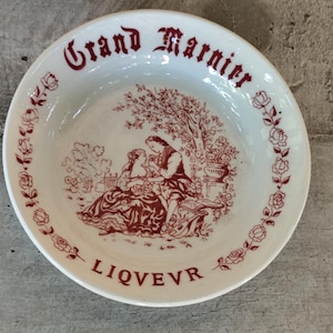 Peut inclure: Bol en céramique blanc avec une illustration et du texte rouges. Le bol présente une scène romantique et les mots "Grand Marnier" et "LIQVEVR". Des motifs floraux et de feuilles ornent le bord.
