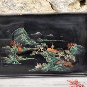 Puede incluir: Bandeja rectangular negra con una escena de paisaje pintada. La obra de arte representa montañas verdes, una pagoda y figuras, con detalles en naranja y blanco. La bandeja tiene un acabado brillante y un aspecto ligeramente desgastado.
