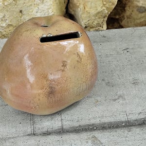 Könnte beinhalten: Ein Keramik-Sparschwein in Form eines Apfels mit einem Schlitz für Münzen. Das Sparschwein ist in einem hellbraunen Ton glasiert und hat eine craquelée-Oberfläche.