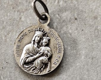 Puede incluir: Un colgante plateado con una imagen religiosa de una mujer coronada sosteniendo a un niño, con el texto "NOTRE DAME DU SACRE COEUR P.P.R." alrededor del borde. El colgante tiene un lazo en la parte superior.