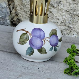 Op de afbeelding: Een witte, ronde keramische geurlamp met een goudkleurige metalen bovenkant. De lamp is versierd met een tak met paarse pruimen en groene bladeren. De lamp staat op een grijze ondergrond.