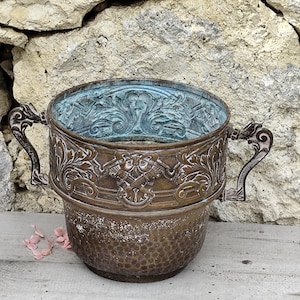 Op de afbeelding: Een antieke bronzen plantenbak met sierlijke handvatten en gedetailleerde reliëfpatronen. De plantenbak heeft een getextureerde, gehamerde afwerking en een verweerde patina, met een turquoisekleurig interieur. De handvatten hebben decoratieve krullen.