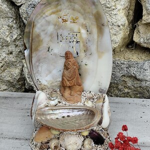 Puede incluir: Una pequeña figurita de arcilla de una mujer con un chal se exhibe dentro de una gran concha de nácar iridiscente. La concha está rodeada de conchas más pequeñas y se asienta sobre una base hecha de arena y conchas. La base está decorada con una variedad de conchas, incluida una gran concha de vieira blanca.