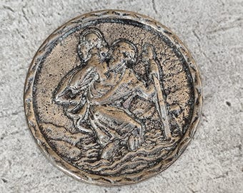 May include: A round, bronze-toned metal button featuring a raised relief design of a figure carrying a child on their shoulder while holding a tall staff. The surface has a textured, weathered finish with dark oxidation in the recessed areas.