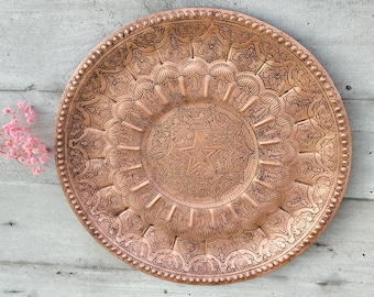 May include: A large, round, copper serving tray with intricate embossed designs. The tray features a central starburst pattern surrounded by a ring of smaller, repeating motifs.