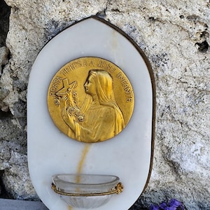 May include: A religious wall hanging with a gold medallion inscribed "BEATA TERESIA A JESU INFANTE" and a woman's image. The medallion is set on a white, oval plaque with a small glass bowl at the base. The plaque has a brass-colored frame.