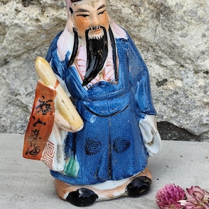 Peut inclure: Figurine en céramique d'un homme asiatique en robe bleue, tenant un rouleau. La figurine a une longue barbe noire et un chapeau. Le rouleau porte des caractères chinois. La figurine est sur une surface grise.