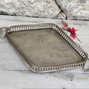 Op de afbeelding: Een antiek zilverkleurig dienblad met sierlijke handgrepen en een decoratieve rand. De lade heeft een ruitvorm en ligt op een verweerd houten oppervlak. Een takje rode bloemen ligt op de lade.