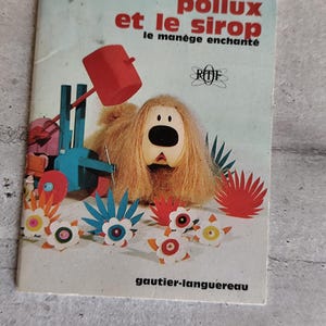 Puede incluir: Una portada de libro vintage titulada "Pollux et le sirop le manège enchanté" con un personaje de perro de dibujos animados con pelo largo, lanudo y marrón claro. La portada tiene un esquema de color rojo y azul con diseños florales y geométricos.