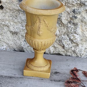Könnte beinhalten: Eine gelbliche, dekorative Urne mit quadratischem Sockel und geriffelten Details. Die Urne weist florale und bandförmige Reliefmuster am Körper auf. Der obere Rand hat eine gerippte Textur. Ein klassisches Stück für die Inneneinrichtung.