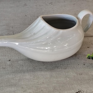 May include: A white ceramic neti pot with a curved spout and a handle. The pot has a smooth, glossy finish and a rounded body. The neti pot is designed for nasal irrigation and sits on a gray surface.
