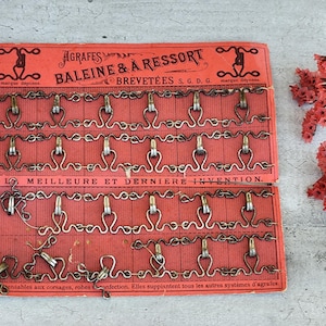 Peut inclure: Une carte rouge avec une bordure blanche et des lettres dorées. La carte fait la publicité d'« Agrafes Baleine & A Ressort », un type de fermoir à ressort. La carte présente un motif de fermoirs en rangées. Le texte sur la carte indique « Marque Deposee » et « Brevetees S.G.D.G. ».