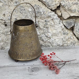 May include: A small, brass bucket with a handle and intricate engraved designs. The bucket is sitting on a wooden surface with a sprig of red flowers.