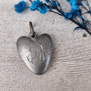 May include: A silver-toned heart-shaped pendant with a loop for a chain. The pendant features an engraved initial "B" in an ornate script. Blue flowers are in the background.