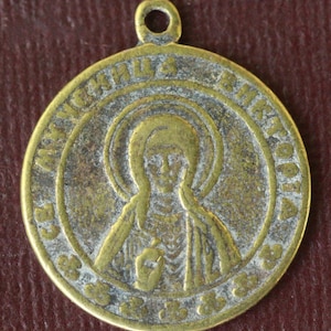 Könnte beinhalten: Ein goldfarbener Metallanhänger mit einem religiösen Symbol einer Frau in einem Heiligenschein. Der Anhänger trägt eine Inschrift am Rand, die lautet "Св. АНАСТАСИЯ В ИКОТОРИЯ".