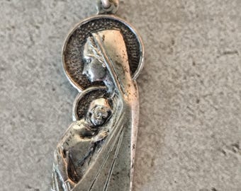 Puede incluir: Colgante de plata con motivo religioso. Representa a una mujer con halo sosteniendo a un niño. El diseño es detallado, con rasgos faciales y pliegues de ropa visibles. Tiene un pequeño lazo en la parte superior para sujetarlo.