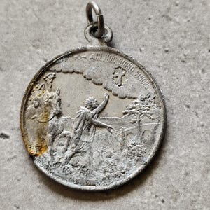 Peut inclure: Un pendentif rond argenté avec une scène en relief détaillée. Le pendentif représente une figure à cheval, une personne pointant vers le haut et l'inscription "IN HOC SIGNO VINCIS". Une petite boucle est en haut pour une chaîne.