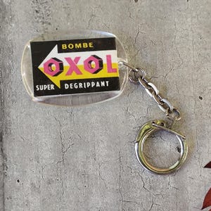 Könnte beinhalten: Ein klarer, ovaler Schlüsselanhänger mit einer Metallkette und einem Ring. Der Schlüsselanhänger hat einen rechteckigen Einsatz mit dem Text "BOMBE OXOL SUPER DEGRIPPANT" in Schwarz, Gelb und Rosa. Der Hintergrund ist eine graue, strukturierte Oberfläche.