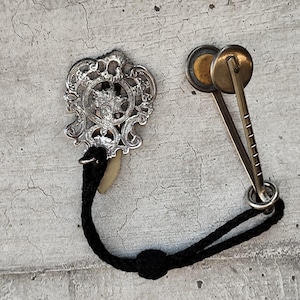 Pode incluir: Um broche de metal prateado com um design ornamentado em forma de escudo com volutas intrincadas, ligado por um cordão trançado preto a um par de clipes circulares de metal latonado com um mecanismo de dobradiça dentado para fixação.